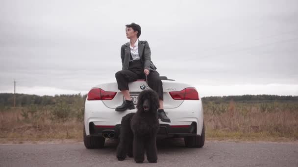 Minsk, Belarus- 23 de setembro de 2021: A jovem morena espetacular com cabelo curto segura um poodle preto em uma coleira enquanto se senta no tronco de um conversível em uma estrada rural. Movimento lento — Vídeo de Stock