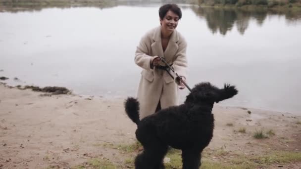 Stylish young woman in a white coat enjoys a walk by the river and plays with a large black poodle. Slow motion — Stock Video