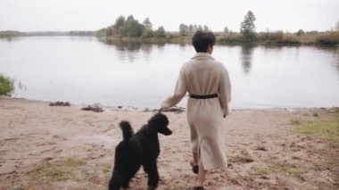 Beyaz önlüklü genç bir esmer nehre gider ve tasmalı siyah bir kaniş tutar. Arkadan bak. Yavaş çekim