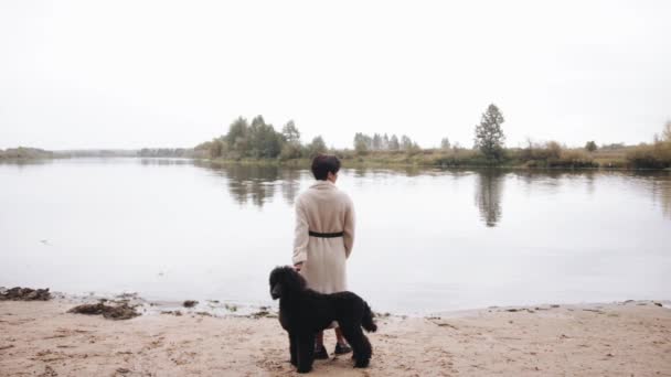 A young woman in a stylish white coat stands by the river with her black poodle and enjoys the autumn landscape. Back view. Slow motion — Stock Video
