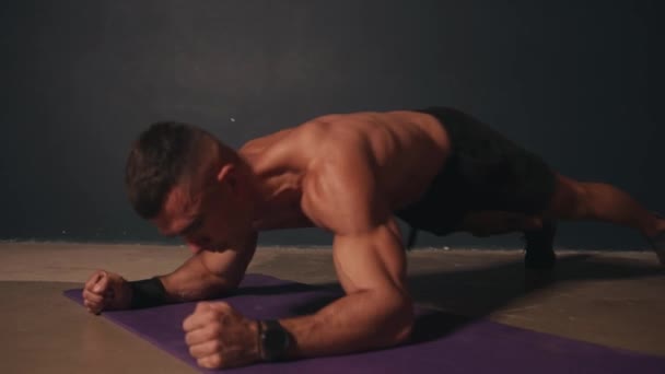 Jonge atletische man in zwarte shorts is training in de kamer op het tapijt en het doen van de plank oefening op de ellebogen met de beweging van armen en benen — Stockvideo