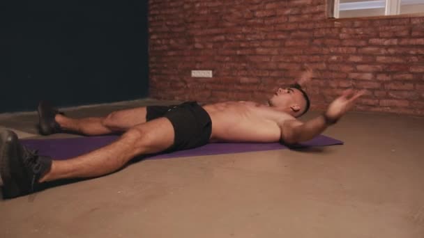 Mladý svalnatý muž školení v místnosti a dělá břišní cvičení tím, že se dotýká nohou s rukama, zatímco leží na zádech na podložce — Stock video