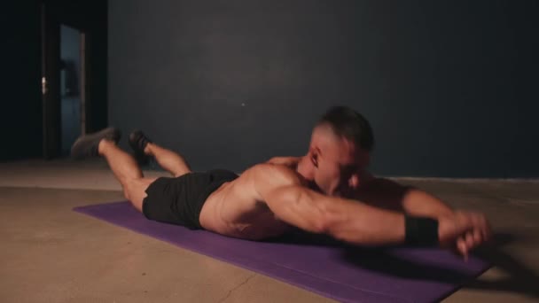 Молодой мускулистый мужчина с голым туловищем тренируется в комнате и делает упражнения супермена лежа на коврике и раскачивания — стоковое видео