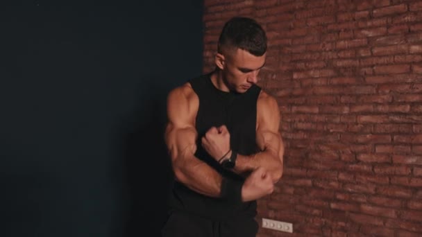 Mladý svalnatý muž v černém tričku bez rukávů stojí na pozadí červené cihlové zdi a demonstruje velké biceps — Stock video