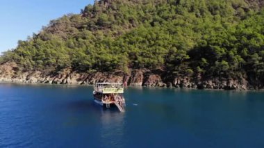 Türkiye, Kemer - 20 Ekim 2021: Turistlerle güneşlenen ve ağaçlarla kaplı kayalık bir adanın yakınlarındaki berrak mavi sularda yüzen bir yatın insansız hava aracı vuruşu
