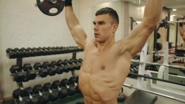 Genç kaslı adam spor salonunda antrenman yapıyor ve halterle ağırlık kaldırıyor. Omuz kemerinin kaslarını geliştirmek için bankta oturuyor.