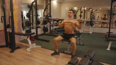 Spor salonunda antrenman yapan genç bir vücut geliştirici. Göğüs kafesinden halter presi yapıyor. Deltoid ve trisepsi pompalarken oturuyor.
