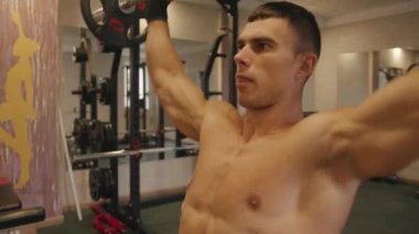 Çıplak gövdeli genç bir adam spor salonunda antrenman yapıyor ve kendine çabalarken barı kaldırıyor.