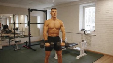 Genç kaslı adam spor salonunda antrenman yapıyor ve önünde duran dambıllarla düz kollar kaldırarak kaslarını çalıştırıyor.