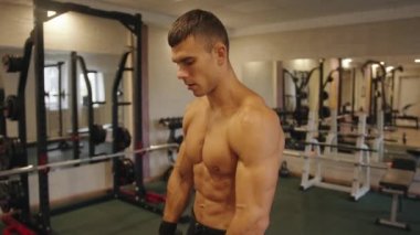 Vücut geliştirici, spor salonunda çıplak gövde eğitimi alıyor ve dambıllarla kol egzersizleri yapıyor.