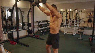 Genç kaslı adam spor salonunda çalışıyor ve kaslarını çalıştırmak için dambıllarla düz kollar sallıyor.