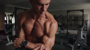 Spordan sonra çıplak gövdeli genç bir adam spor salonunda duruyor ve kolların ve omuz kemerinin şişmiş kaslarını gösteriyor.