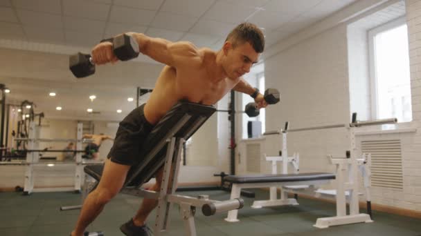 Mladý svalnatý muž s nahým trupem cvičí v tělocvičně a zvedá paže s činkami ležícími na lavičce čelem dolů — Stock video