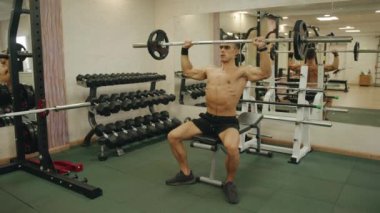 Genç kaslı adam spor salonunda antrenman yapıyor ve halterle ağırlık kaldırıyor. Omuz kemerinin kaslarını geliştirmek için bankta oturuyor.