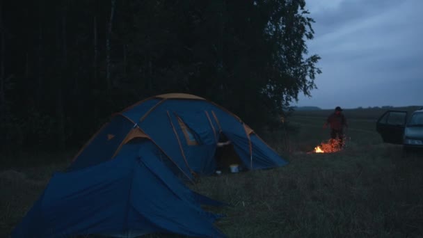 Um jovem em um boné sai de uma tenda em um acampamento nos arredores da floresta ao lado de uma fogueira e um carro estacionado — Vídeo de Stock