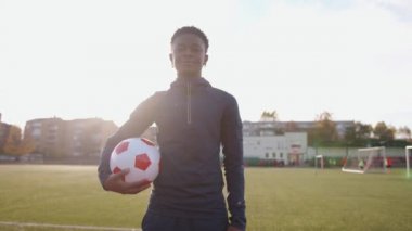 Spor üniformalı genç siyah bir kız stadyumda duruyor ve elinde bir futbol topu tutuyor. Yavaş çekim