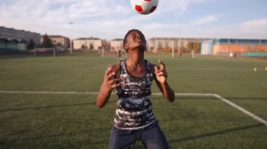 Kolsuz tişörtlü ve şortlu Afro-Amerikalı genç kız stadyumda egzersiz yapıyor ve kafasıyla futbol topuna vuruyor.