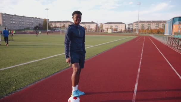 Een jong zwart meisje staat op een stadion loopband met haar voet op een voetbal en wijst er met haar wijsvinger naar. Langzame beweging — Stockvideo