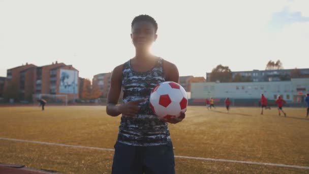 Молода чорна дівчина стоїть на стадіоні і тримає в руці футбольний м'яч і вказує на нього пальцем іншої руки. Повільний рух — стокове відео