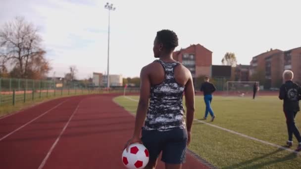 En ung afroamerikansk kvinna i ärmlös skjorta går på ett stadion löpband med en fotboll i handen. Bakåt. Långsamma rörelser — Stockvideo