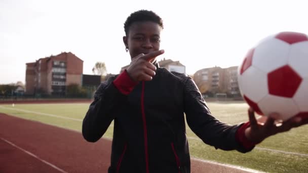 Młoda piękna czarna dziewczyna stoi na stadionie trzymając piłkę nożną i wskazuje na to i pokazuje kciuk w górę. Zwolniony ruch — Wideo stockowe
