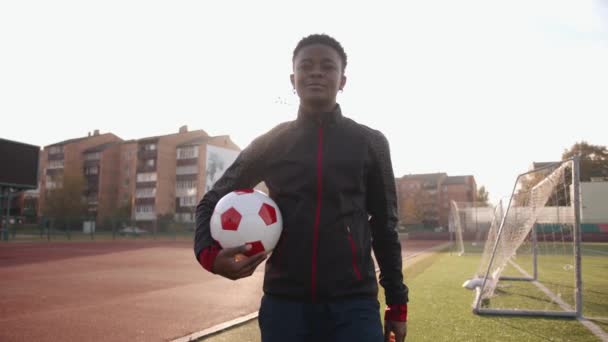 Una joven negra enérgica con uniforme deportivo cruza el campo de fútbol con una pelota en las manos y sonríe. Movimiento lento — Vídeos de Stock