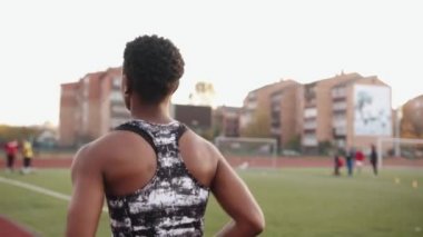 Kolsuz tişörtlü genç siyah bir kız stadyuma spor yapmaya gider ve koşu bandında koşar. Arkadan bak. Yavaş çekim