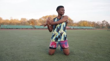 Genç siyah kız parlak spor üniforması futbol sahasında antrenman yapıyor ve diz çökerken kollarını esnetiyor.