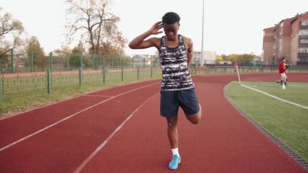 Una joven negra va a practicar deportes en la cinta de correr del estadio y hace ejercicios de estiramiento estáticos. Movimiento lento — Vídeos de Stock