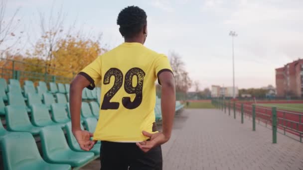 Młoda Afroamerykanka stoi w koszulce piłkarskiej na stadionie i wskazuje dwa kciuki na numer na plecach. Zwolniony ruch — Wideo stockowe