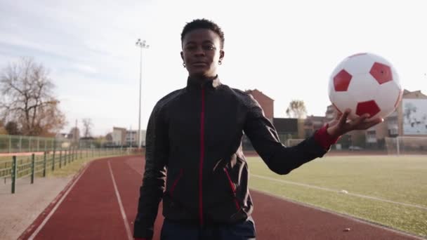 若いアフリカ系アメリカ人の少女が太陽の光の中のスタジアムに立っていて、手にサッカーボールを持っている。スローモーション — ストック動画