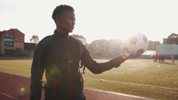 Una giovane ragazza afroamericana si trova in uno stadio della città al sole e tiene un pallone da calcio sulla mano tesa. Rallentatore — Video Stock