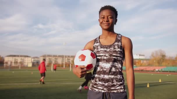 Ein fröhliches schwarzes Mädchen steht mit einem Fußballball in der Hand vor dem Hintergrund des Mannschaftstrainings im Stadion. Zeitlupe — Stockvideo