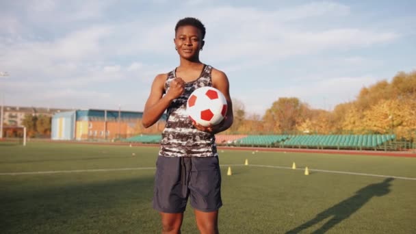 Весела молода чорна дівчина стоїть з футбольним м'ячем в руках на тлі командного тренування на стадіоні. Повільний рух — стокове відео