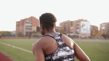 Kolsuz tişörtlü genç siyah bir kız stadyuma spor yapmaya gider ve koşu bandında koşar. Arkadan bak. Yavaş çekim