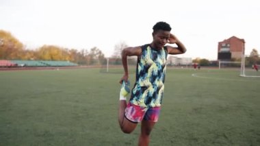Genç Afrikalı Amerikalı kadın futbol sahasında antrenman yapıyor ve tek ayak üstünde ısınıyor.