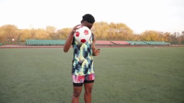 Kolsuz tişörtlü ve şortlu genç bir zenci kız stadyumda duruyor ve yerde bir futbol topuna vuruyor.