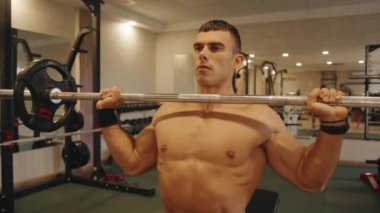 Çıplak gövdeli genç bir adam spor salonunda antrenman yapıyor ve kendine çabalarken barı kaldırıyor.