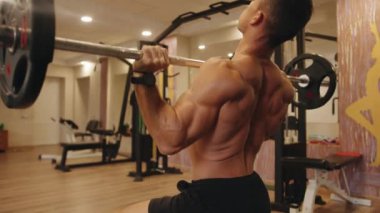 Spor salonundaki genç kaslı bir adamın arkasını yakın çekim. Göğüs kafesinden halterle baskı yapıyor. Bir bankta oturuyor.
