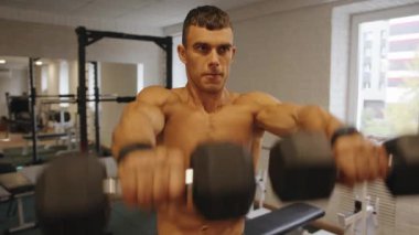 Vücut geliştirici, spor salonunda çıplak gövde eğitimi alıyor ve dambıllarla kol egzersizleri yapıyor.