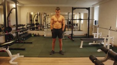 Siyah şortlu ve siyah ayakkabılı genç kaslı adam spor salonunun ortasında elleri cebinde duruyor.
