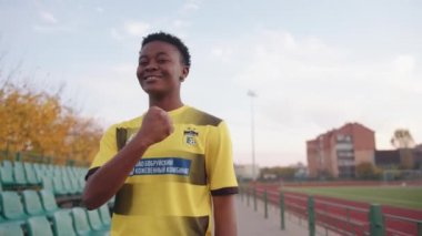 Belarus, Minsk - 20 Haziran 2021: Genç siyah bir kız futbol takımının üniformasıyla stadyumda duruyor ve sıkılmış yumruğunu gülümserken gösteriyor. Yavaş çekim