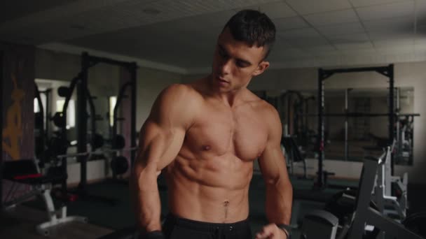 Un culturista sin camiseta muestra sus músculos bombeados mientras está de pie en el gimnasio y se golpea el bíceps con los puños — Vídeo de stock