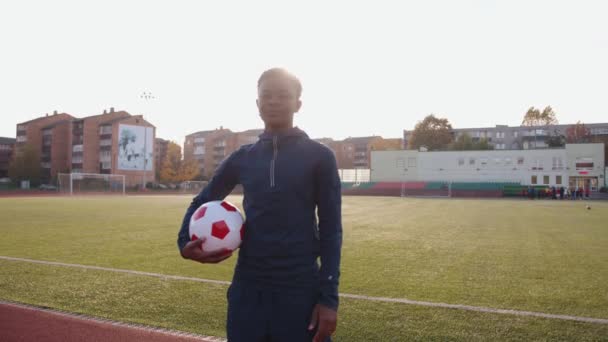 Een energiek jong zwart meisje in een sportuniform gaat over het voetbalveld met een bal in haar handen en glimlacht. Langzame beweging — Stockvideo