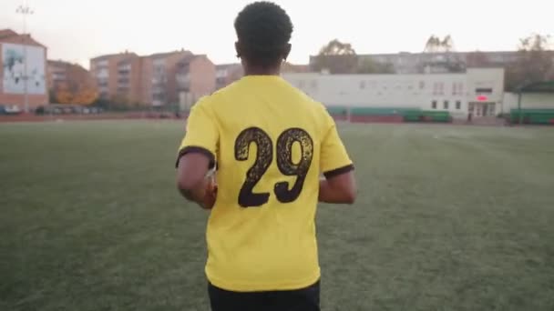 Jovem negro enérgico de uniforme de futebol com um número na parte de trás passa pelo estádio e olha para a câmera — Vídeo de Stock