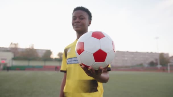 Улыбающаяся молодая черная девушка стоит на городском стадионе в своей футбольной форме и держит мяч протянутым. Медленное движение — стоковое видео