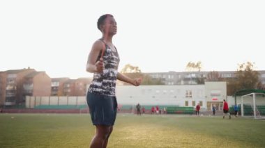 Şehir bloğunda bir futbol sahasında antrenman yapan genç siyah bir kız ve güneşin arka planında hızla atlayan bir atlayıcı.