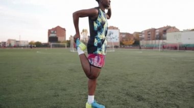 Genç Afrikalı Amerikalı kadın futbol sahasında antrenman yapıyor ve tek ayak üstünde ısınıyor.