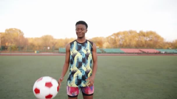 Kolsuz tişörtlü, ciddi bir siyah kız futbol sahasında duruyor ve elinde bir top tutuyor. Yavaş çekim — Stok video