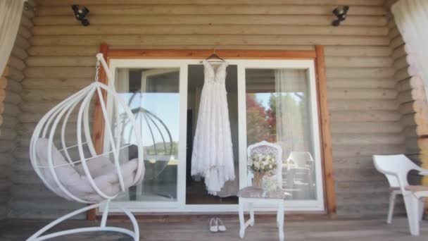 Delikatny strój panny młodej koronki i buty ślubne na balkonie drewnianego domu z huśtawką krzesła i bukietem — Wideo stockowe